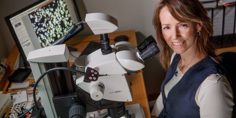 Kvinna ler bredvid ett mikroskop, med en dator i bakgrunden.
