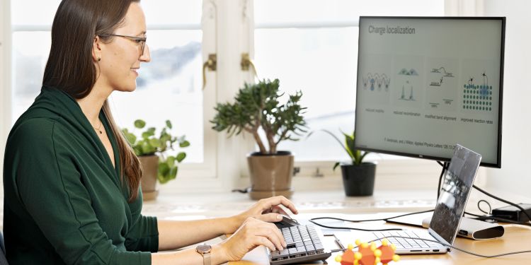 Kvinna arbetar vid dator med grönt plagg, blommor och växter i bakgrunden.