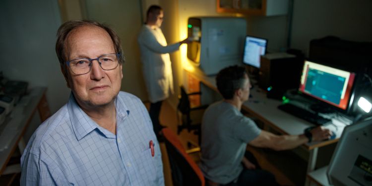 En man står i förgrunden av ett labb, medan två kollegor arbetar vid datorer i bakgrunden.