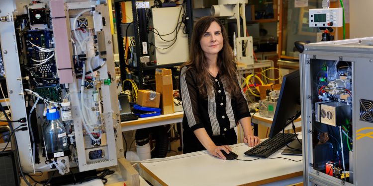 Kvinna står vid ett skrivbord med maskiner och datorutrustning i bakgrunden.