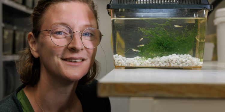 Leende kvinna med glasögon sitter vid ett akvarium med gröna växter och fiskar.