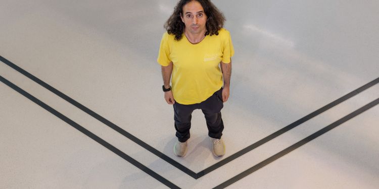 Person med lockigt hår i gul tröja står på en ljus golvyta med svarta linjer.