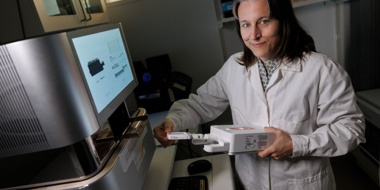 Kvinna i labbrock håller en apparat framför en dator i ett laboratorium.