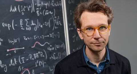 En man med glasögon står framför en svarts tavla med matematiska formler.