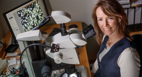 Kvinna ler bredvid ett mikroskop, med en dator i bakgrunden.