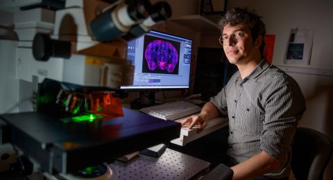 En man sitter vid en dator med en mikroskop i förgrunden, omgiven av forskningsutrustning.