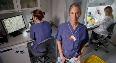 Kvinna i laboratoriemiljö håller en provplatta, andra forskare arbetar i bakgrunden.