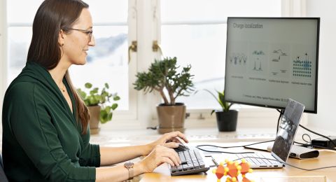 Kvinna arbetar vid dator med grönt plagg, blommor och växter i bakgrunden.