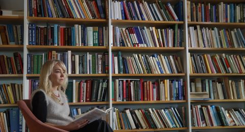 Kvinna sitter i en stol framför en bokhylla med många böcker.