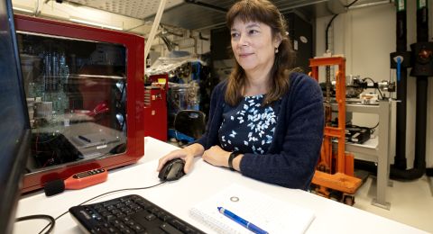 Kvinna sitter vid ett skrivbord framför en dator, omgiven av teknisk utrustning.