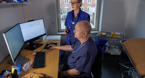 Sjuksköterska står bakom en patient vid dator i ett vårdrum.