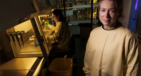 Kvinnlig forskare i ett laboratorium, klädd i skyddsdräkt, ler mot kameran.