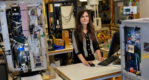 Kvinna står vid ett skrivbord med maskiner och datorutrustning i bakgrunden.