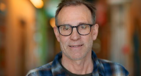 En man med glasögon och rutiga skjortor poserar i en välbelyst korridor.