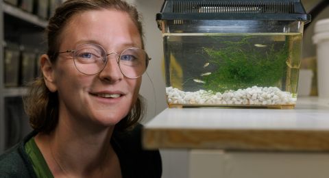 Leende kvinna med glasögon sitter vid ett akvarium med gröna växter och fiskar.