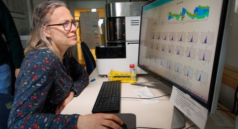 En kvinna med blont hår och prickig skjorta arbetar vid en dator med stora skärmar.