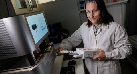 Kvinna i labbrock håller en apparat framför en dator i ett laboratorium.