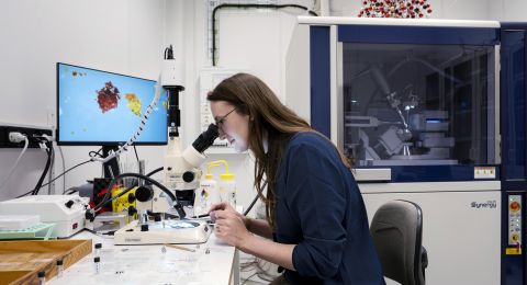 Kvinna som arbetar vid mikroskop i ett laboratorium med en datorskärm i bakgrunden.